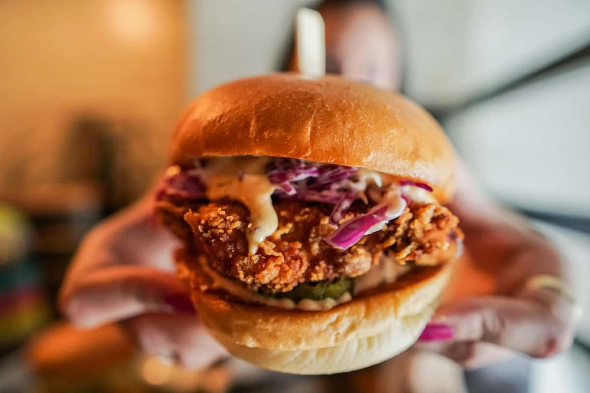 Hand holding a fried chicken sandwich with coleslaw, pickles, and sauce on a toasted brioche bun.
