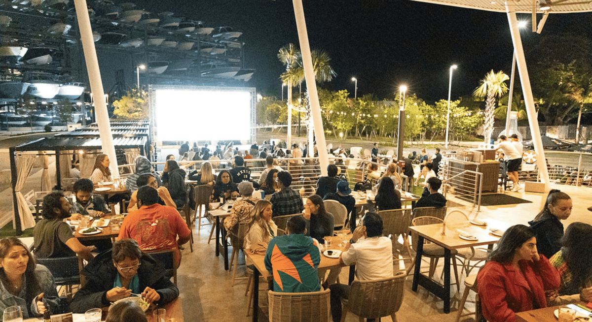 Crowded outdoor waterfront dining space at night with guests seated at tables and a large illuminated screen in view.