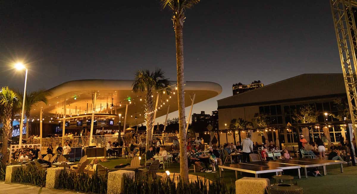 Evening view of a palm-lined waterfront restaurant with illuminated seating areas and people gathering under a modern open canopy.