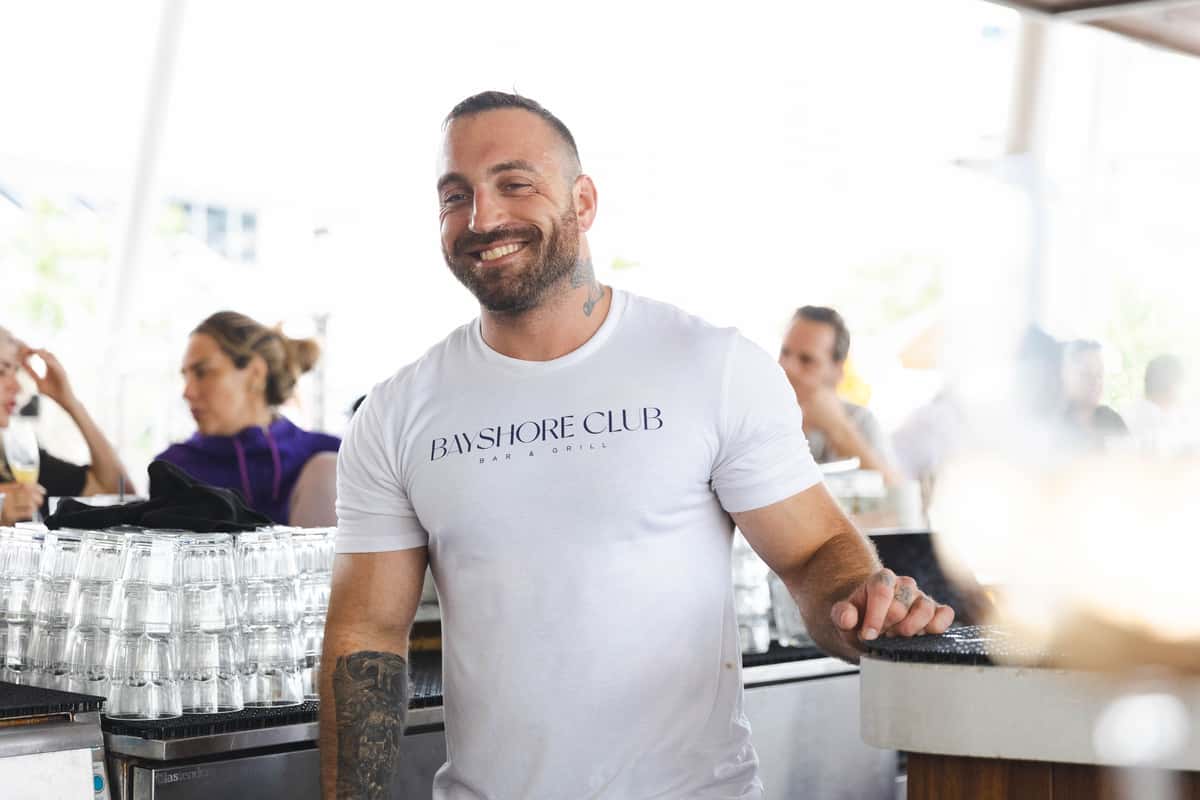 Smiling Bayshore Club bartender standing behind the bar with glassware