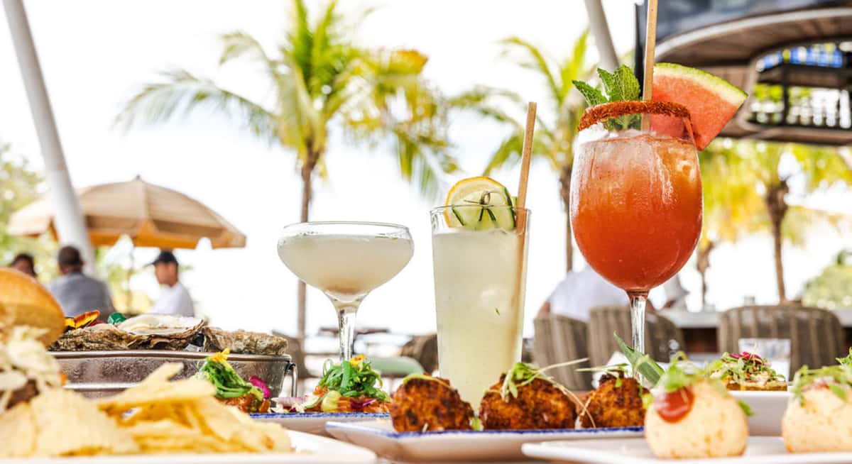 Colorful tropical cocktails and seafood appetizers on a waterfront table with palm trees and outdoor dining in Miami.
