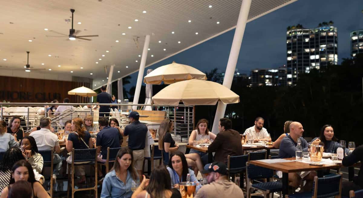 Outdoor waterfront restaurant at night with guests dining under umbrellas and modern lighting overlooking the city skyline.