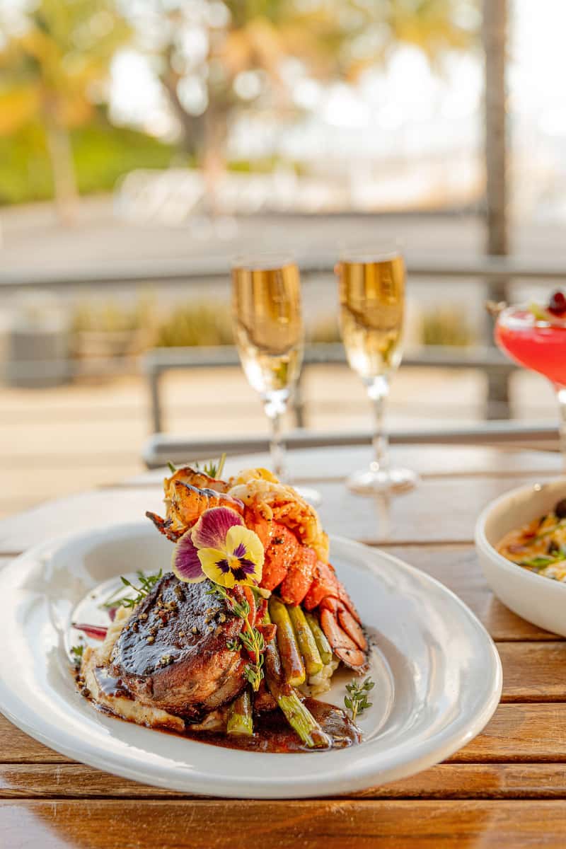 dish.jpg Elegant fine dining dish featuring steak, lobster, asparagus, and edible flowers served on a restaurant table with champagne glasses