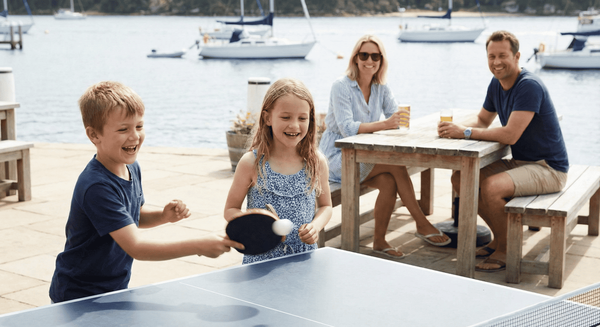 Children playing ping pong at Bayshore Club while families relax by the marina.