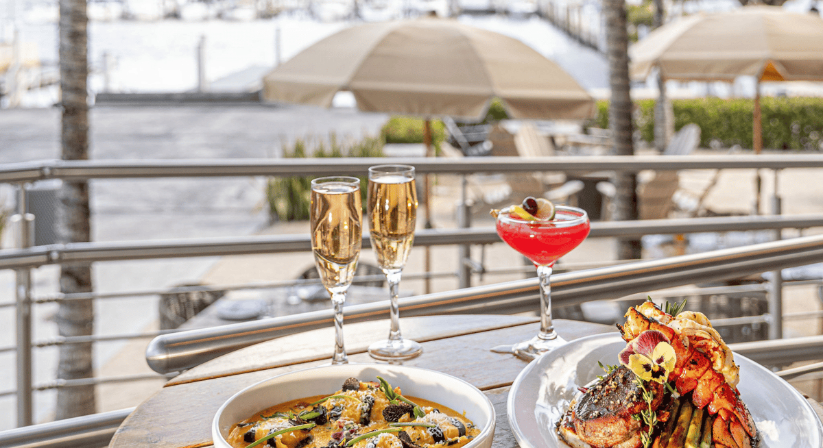 Waterfront restaurant table with champagne flutes, a red cocktail, and plated seafood dishes overlooking a marina patio.