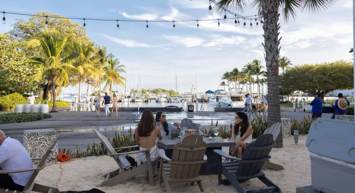 Waterfront dining at Bayshore Club with guests seated outdoors near the marina, overlooking boats, palm trees, and Biscayne Bay.