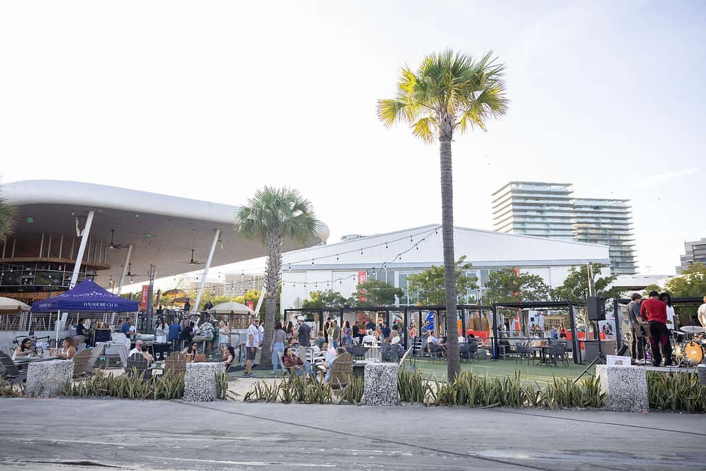Outdoor waterfront restaurant patio with palm trees, seating areas, and guests enjoying a daytime dining event