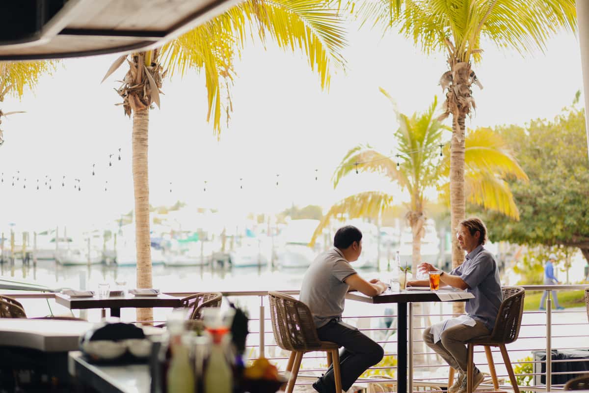 Bayshore Club waterfront dining table with guests seated under palm trees overlooking the marina.