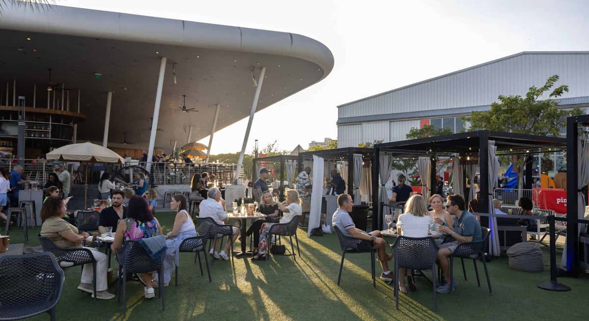 Open-air waterfront dining area with cabanas, tables, and guests enjoying drinks and conversation during golden hour.