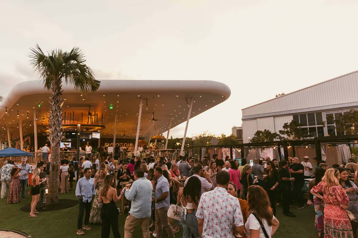 Crowd gathered at an open-air waterfront restaurant during sunset with palm trees, string lights, and views of Biscayne Bay in Coconut Grove, Miami.