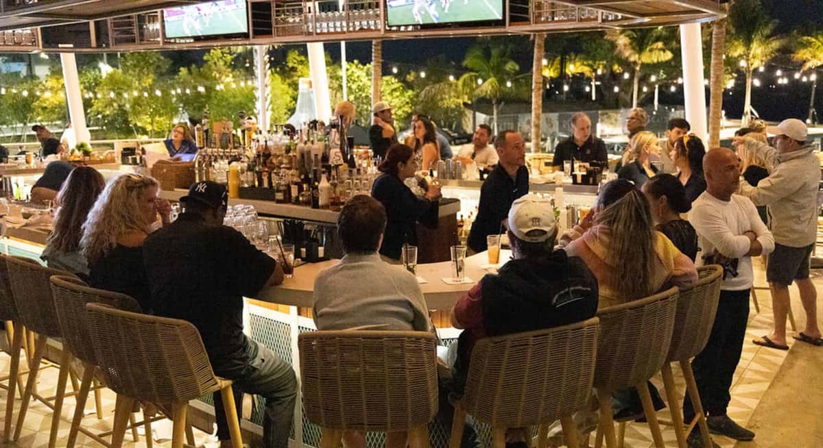 Open-air bar at Bayshore Club in the evening with guests gathered around the counter under string lights and palm trees.