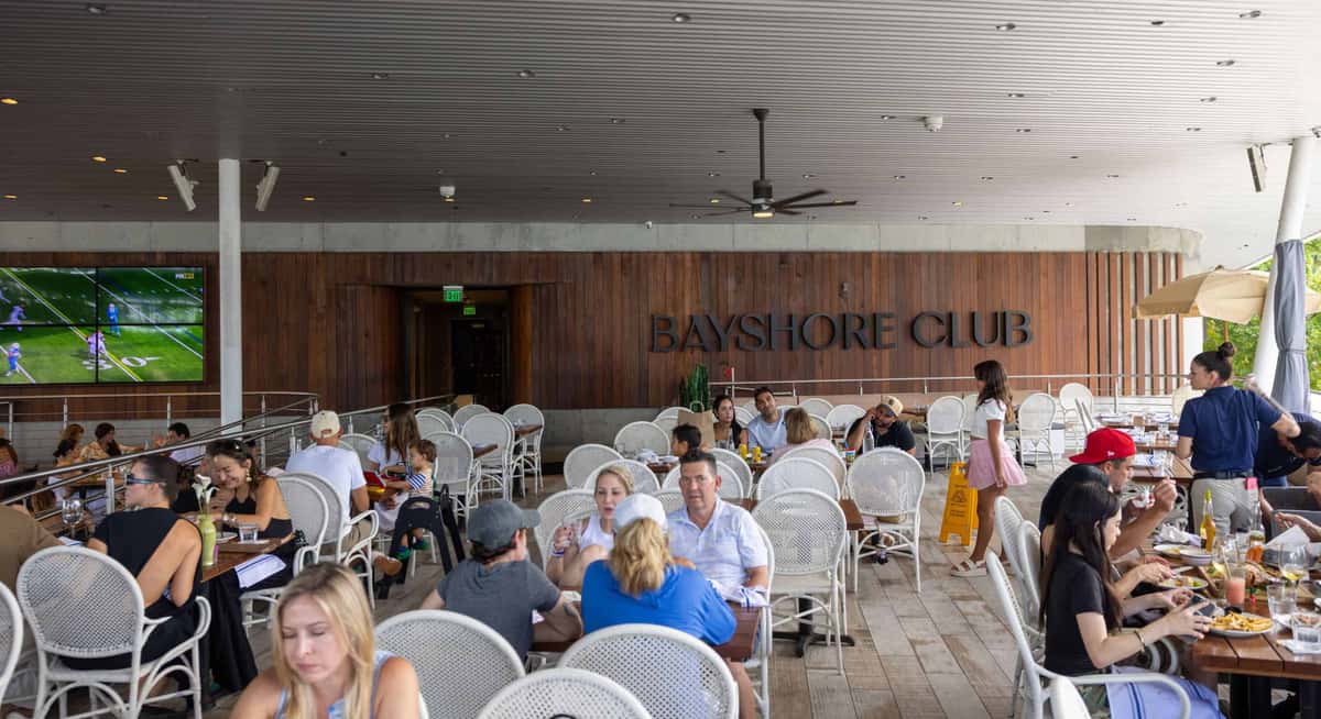 Covered outdoor dining area at Bayshore Club with guests seated at white café tables beneath a modern wood-paneled ceiling.