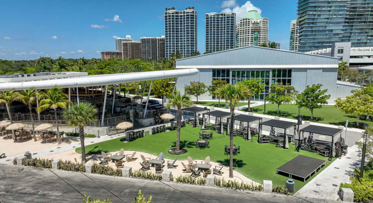 Spacious lawn seating area with private cabanas and palm trees beside a modern waterfront restaurant in Miami.