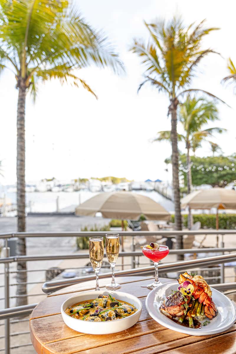 Waterfront dining table with lobster, steak, pasta, champagne, and a cocktail under palm trees