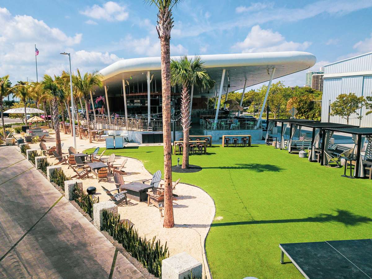 Modern tropical restaurant patio with palm trees, lounge seating, cabanas, and a spacious green lawn.