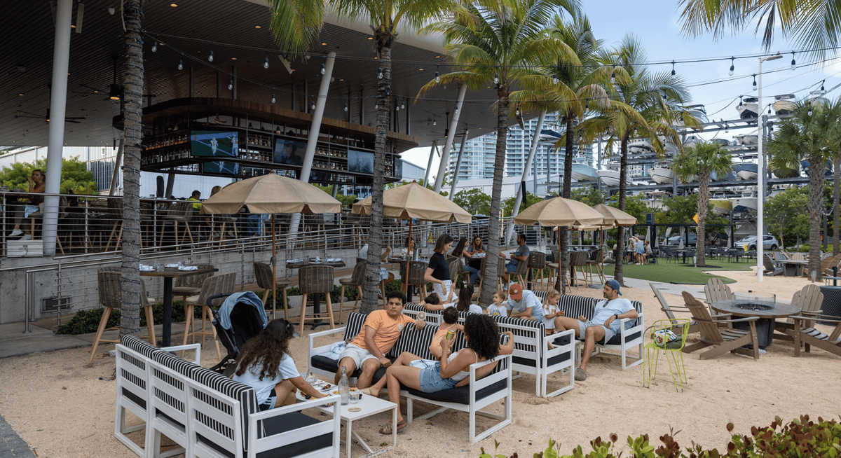 People relaxing in outdoor lounge seating at a waterfront restaurant with palm trees, umbrellas, and a marina nearby.