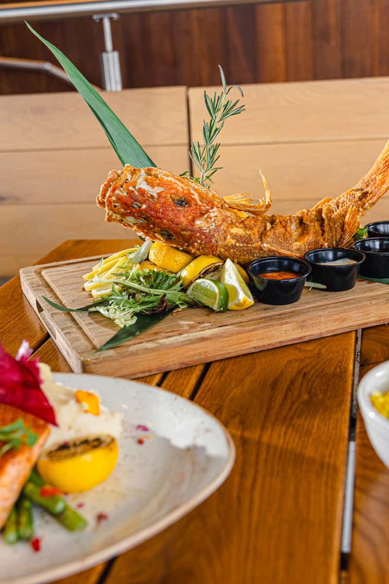 Whole fried fish served on a wooden board with citrus, greens, herbs, and dipping sauces.