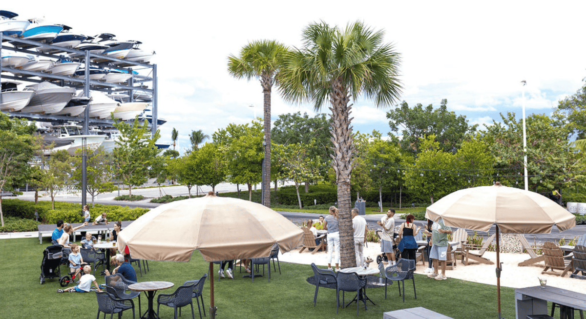 Outdoor waterfront dining area with palm trees, beige umbrellas, and families relaxing on a grassy patio near a marina.