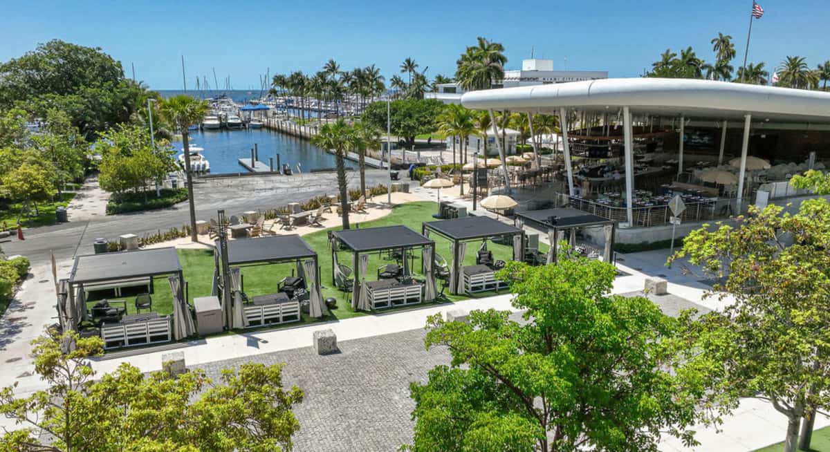 Aerial view of a waterfront restaurant in Coconut Grove with marina boats, palm trees, and outdoor cabanas overlooking Biscayne Bay.