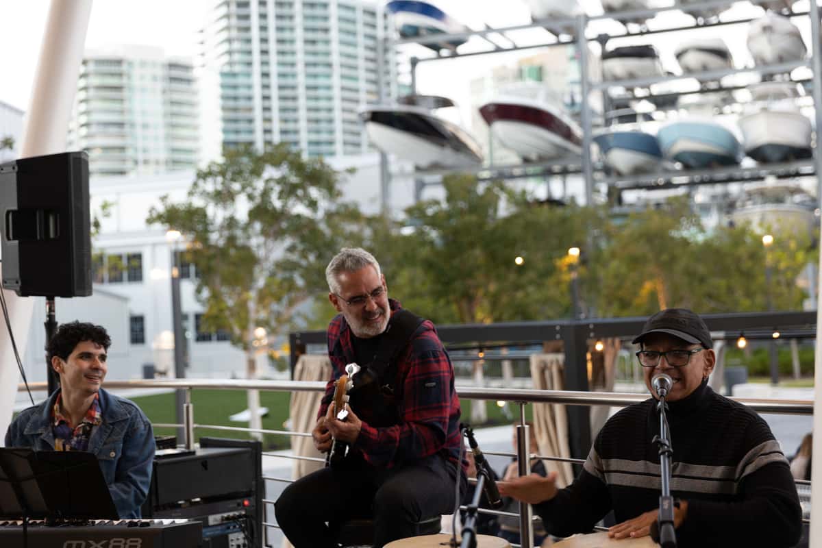 Live band performing at an outdoor waterfront venue with boats and city buildings in the background.