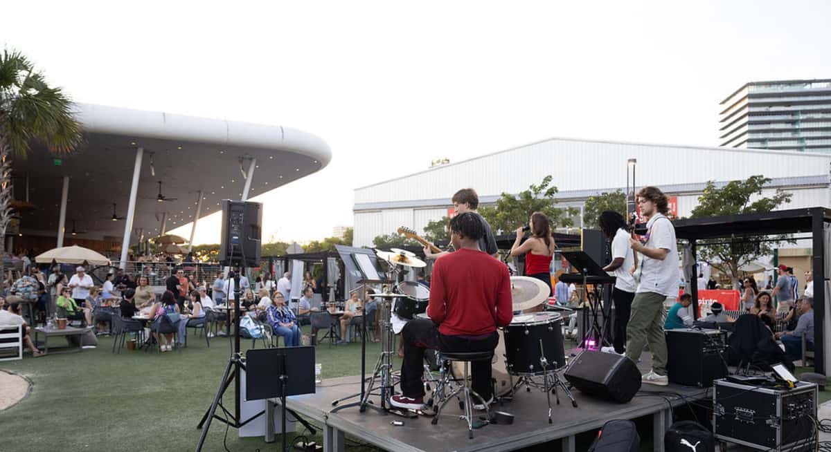 Live band performing on an outdoor stage at a Miami waterfront restaurant with guests seated on a lawn during sunset.
