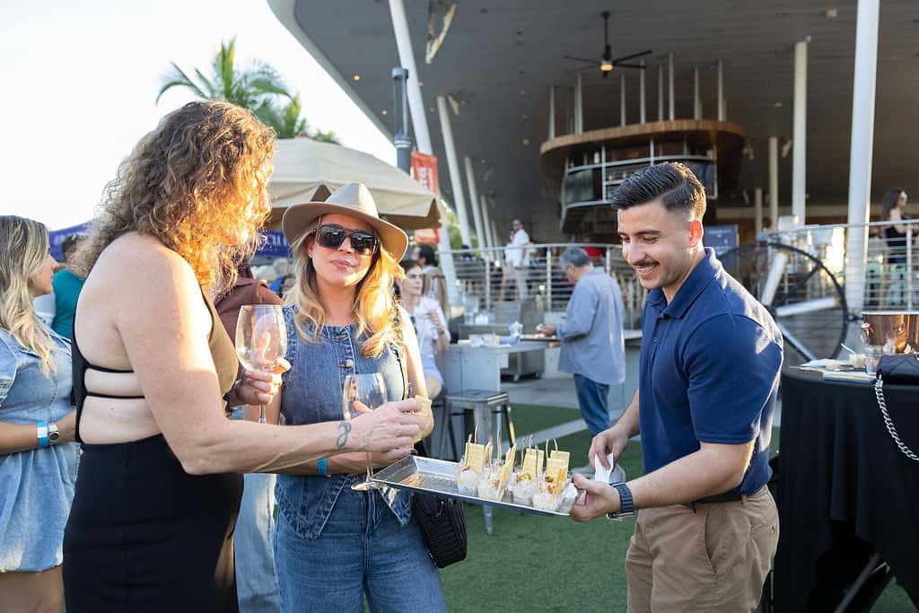Guests enjoying drinks and appetizers at an outdoor restaurant event with a server serving food in a patio setting