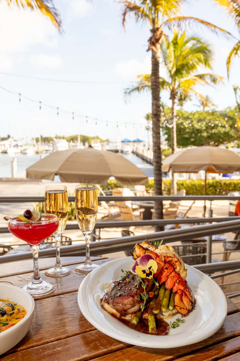 Waterfront fine dining plate with steak, lobster, asparagus, cocktails, and marina views