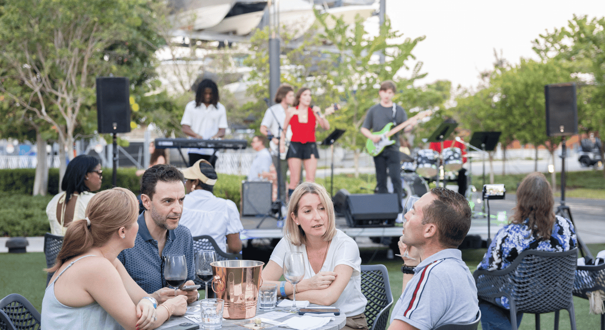 Guests enjoying dinner and wine at an outdoor restaurant while a live band performs on a garden stage.