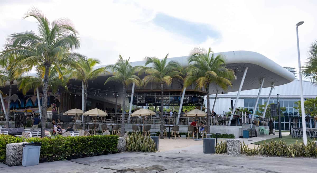 Daytime view of an open-air waterfront restaurant with palm trees, outdoor seating, umbrellas, and a modern curved canopy structure.