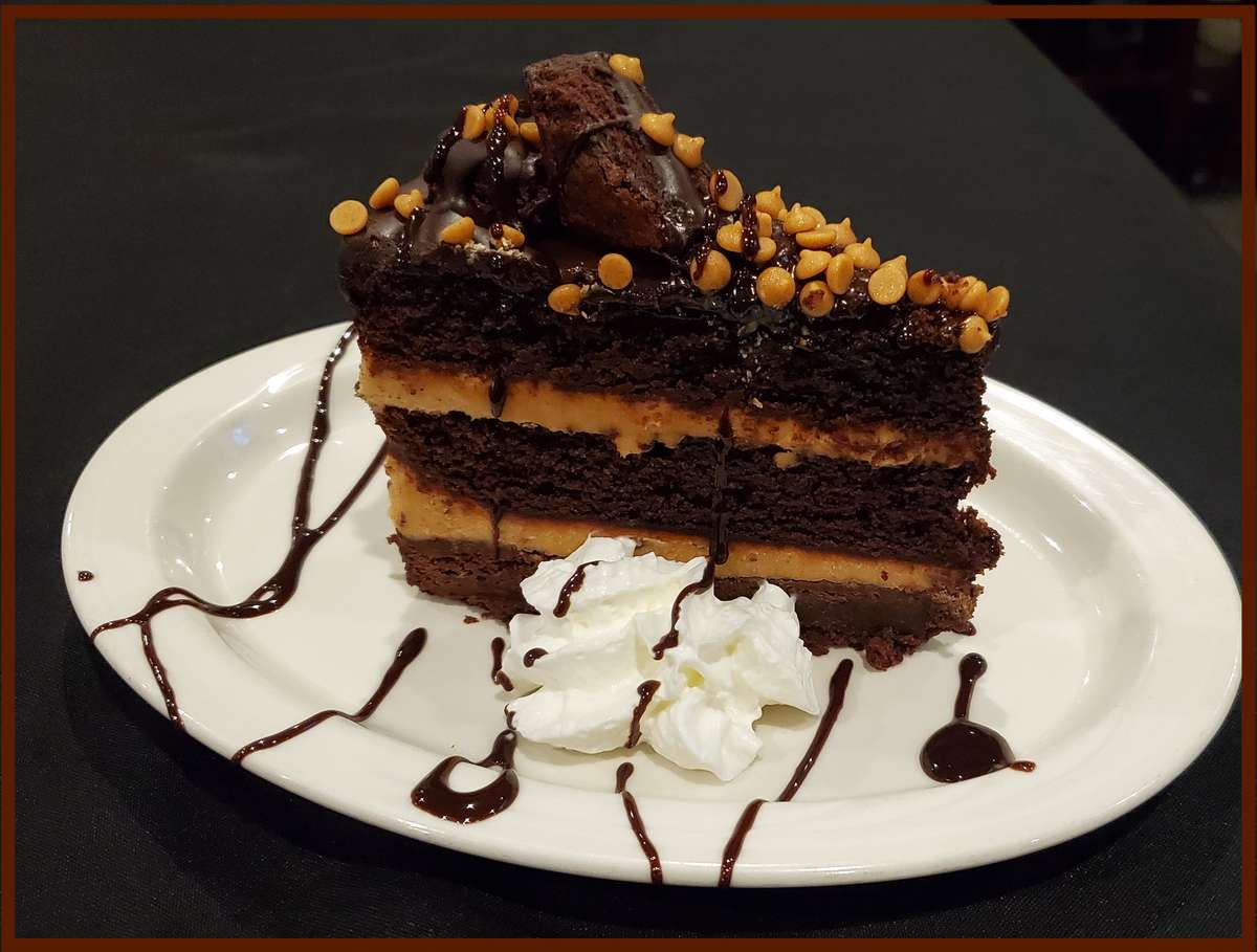 The image showcases a decadent layered chocolate cake topped with crushed nuts and chocolate drizzle. It's served with whipped cream and extra chocolate sauce, making it a rich and indulgent dessert presentation.