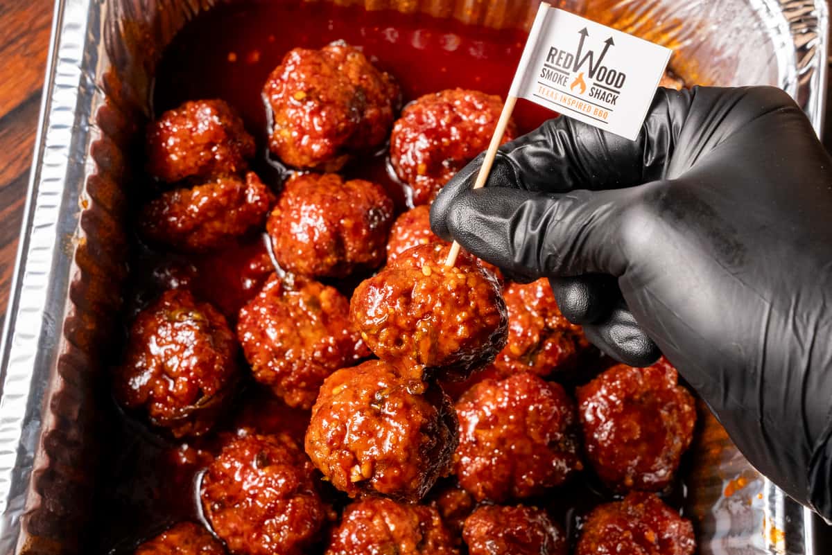 A hand placing a Redwood branded toothpick flag into a catered pan of bbq meatballs