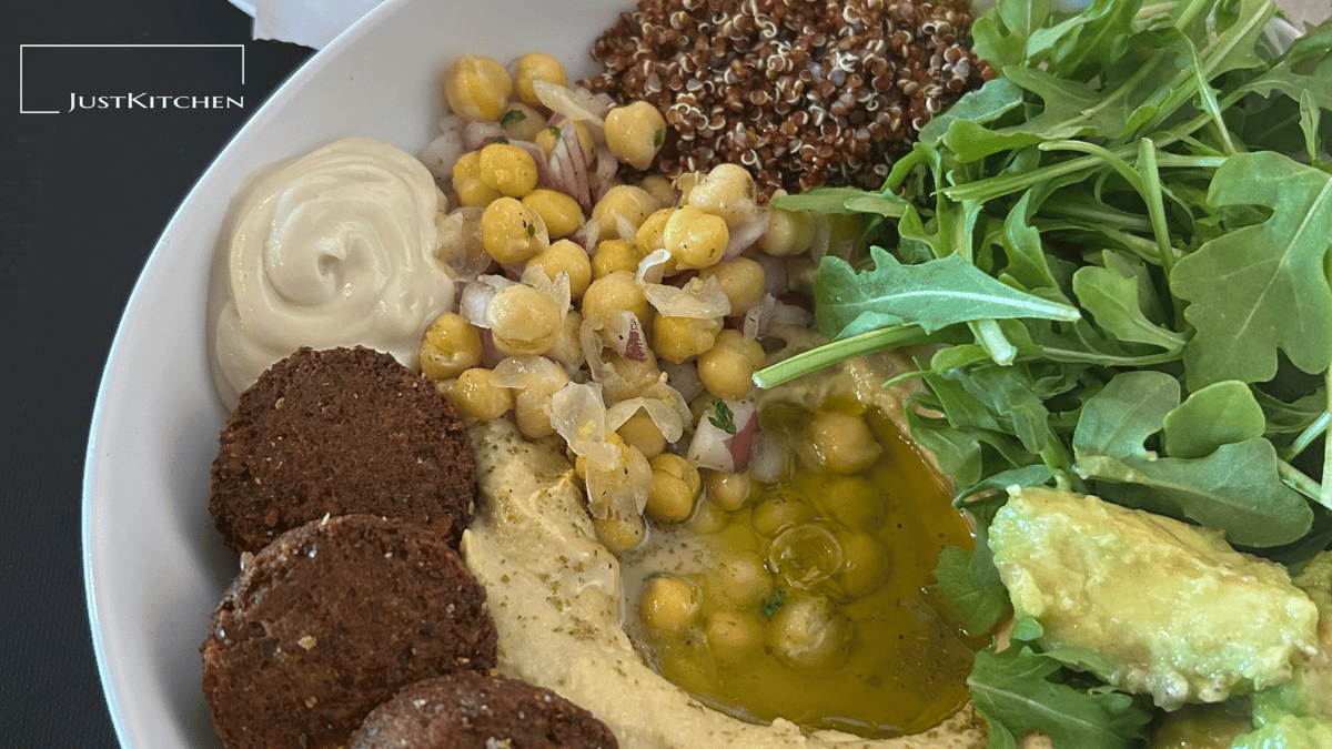 A fresh Mediterranean bowl from Just Kitchen Jax featuring falafel, hummus drizzled with olive oil, chickpeas, quinoa, creamy sauce, avocado, and arugula greens.