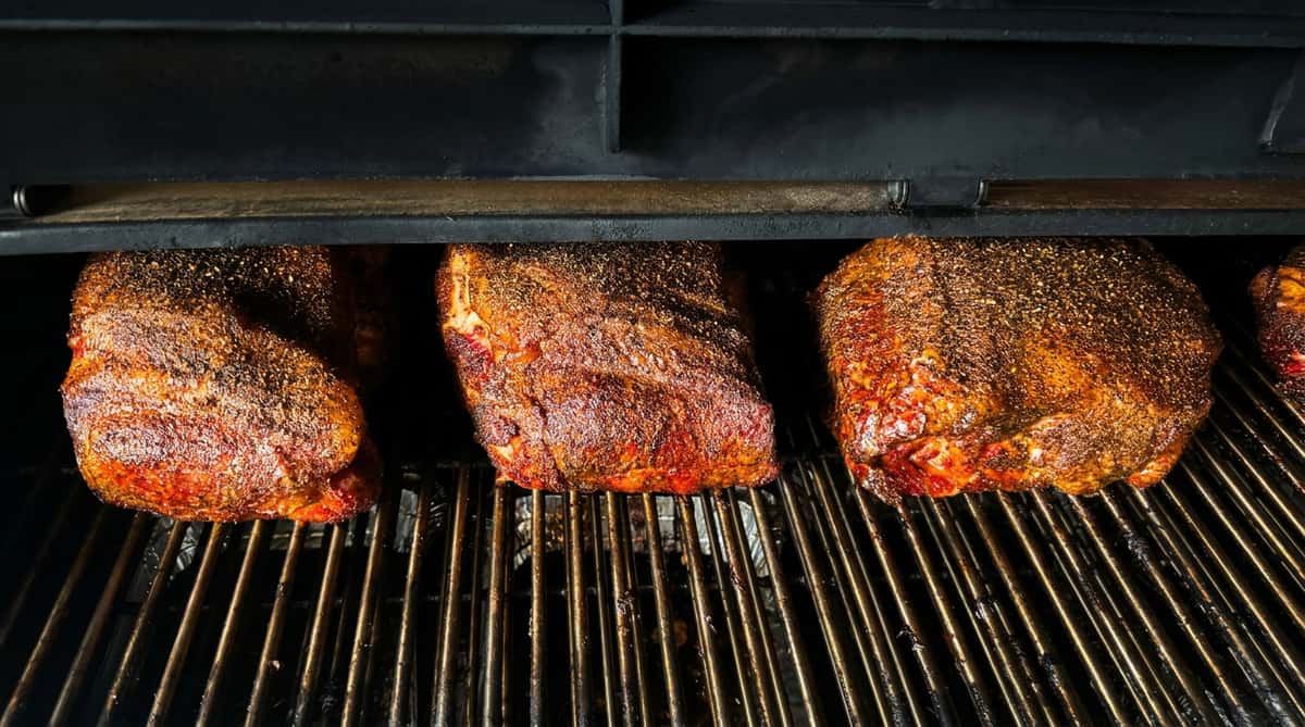 Three large cuts of BBQ meat, seasoned with black pepper and spices, cooking on a dark metal grill inside a smoker.