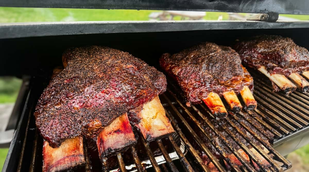 Smoked beef short ribs with a thick pepper rub cooking on a grill, bones visible.