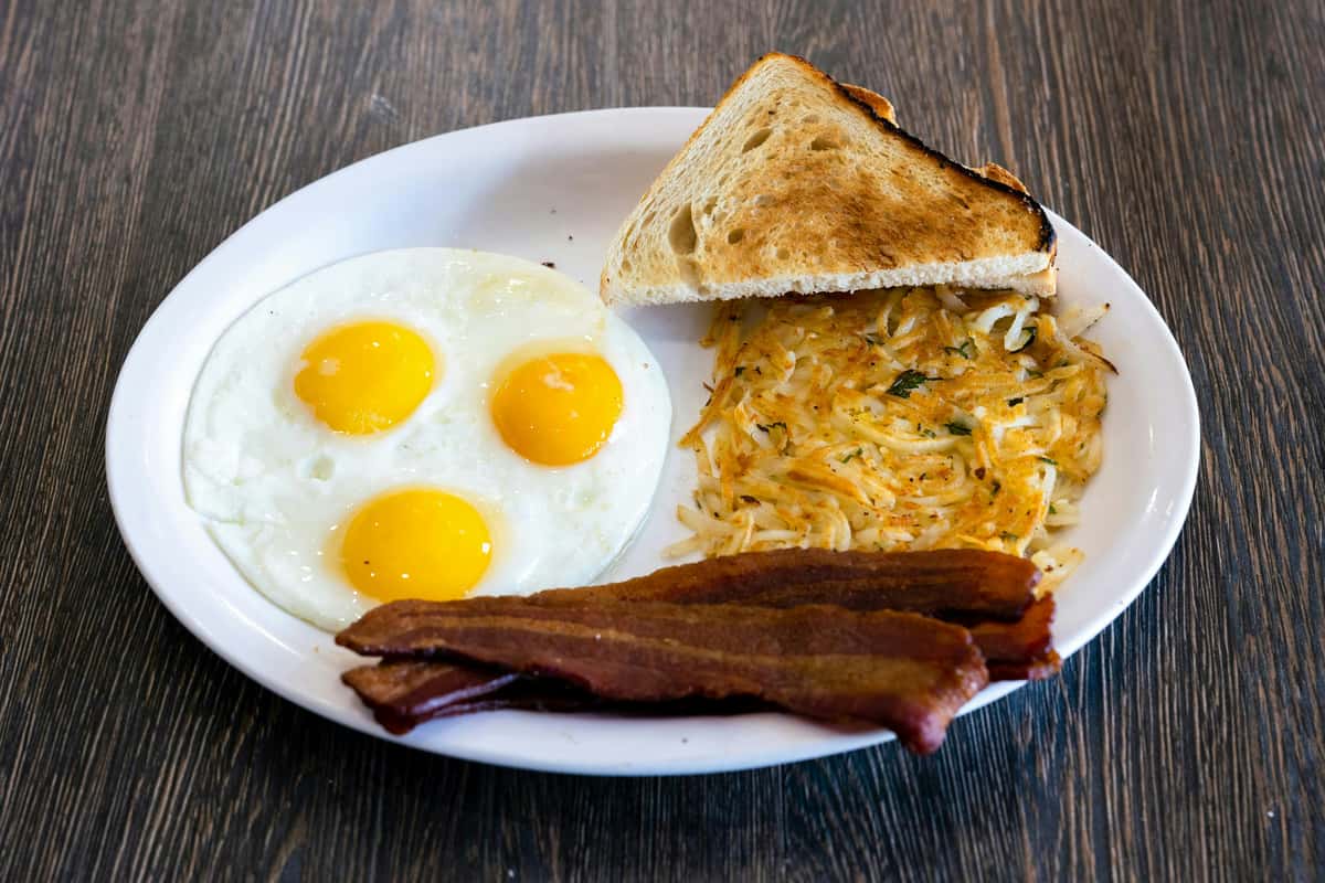 Three fried eggs with crispy bacon strips, golden hash browns, and toasted bread—homestyle breakfast special at a local café.