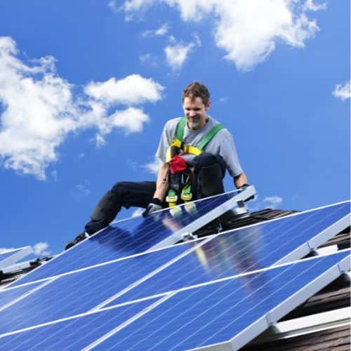 Smiling worker in safety harness installs solar panels on a sloped roof under a bright blue sky with scattered clouds.