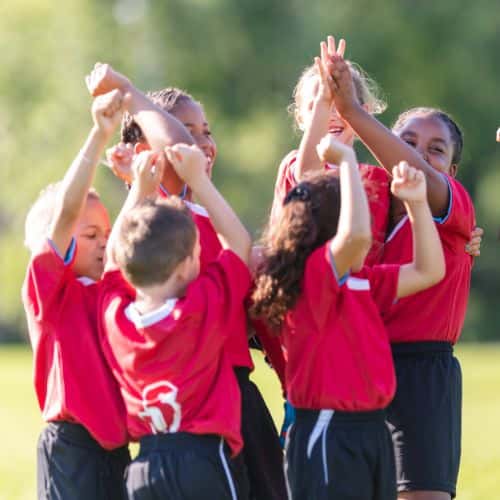 Group of young soccer players in red jerseys huddle together outdoors, raising their arms in celebration and teamwork.