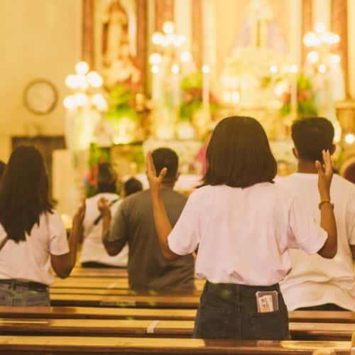People in white shirts stand in wooden pews of a warmly lit church, raising their hands in worship facing the ornate altar.