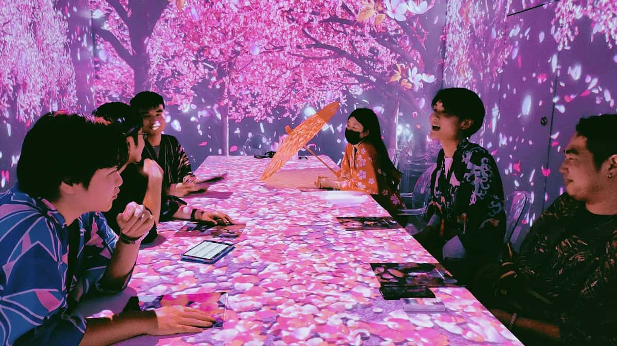 Group of people in floral attire enjoying a cherry blossom-themed room with pink petal projections covering walls and table.