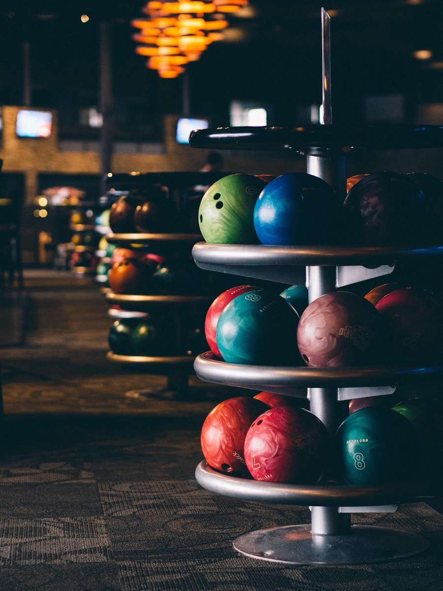 Colorful bowling balls arranged on a metal rack in a dimly lit bowling alley with warm overhead lights.