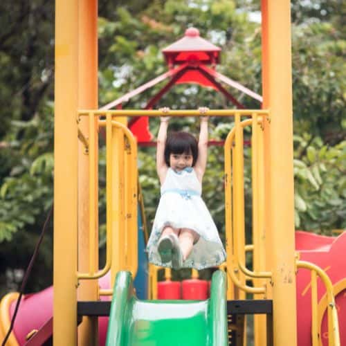 Smiling young girl in a white dress hangs from playground bars above a green slide on a colorful outdoor play structure.