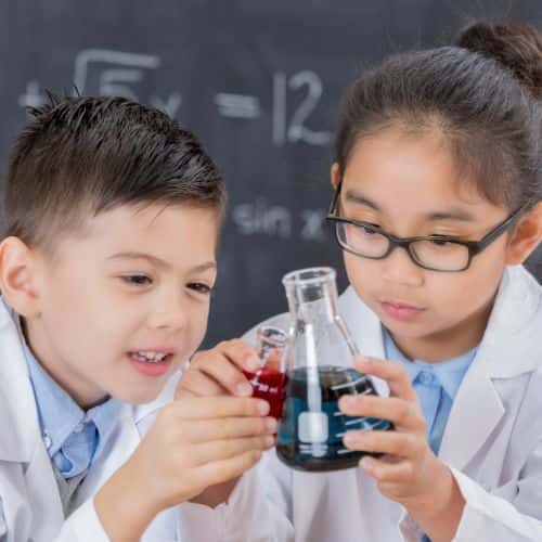 Two children in lab coats examine beakers filled with red and blue liquids in a classroom with math equations on the chalkboard.