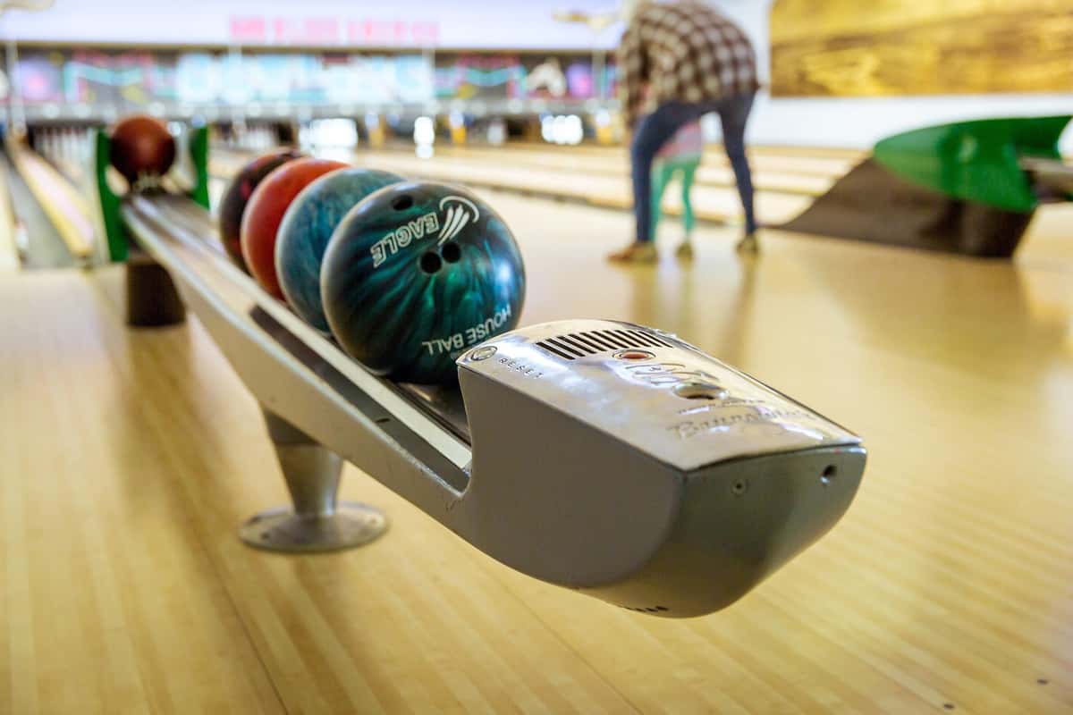 The image shows a close-up of a bowling ball return system with several bowling balls lined up, ready for use. In the background, a person is helping a child aim and roll a ball down the bowling lane. The focus is on the bowling balls and return mechanism, while the background is slightly blurred.