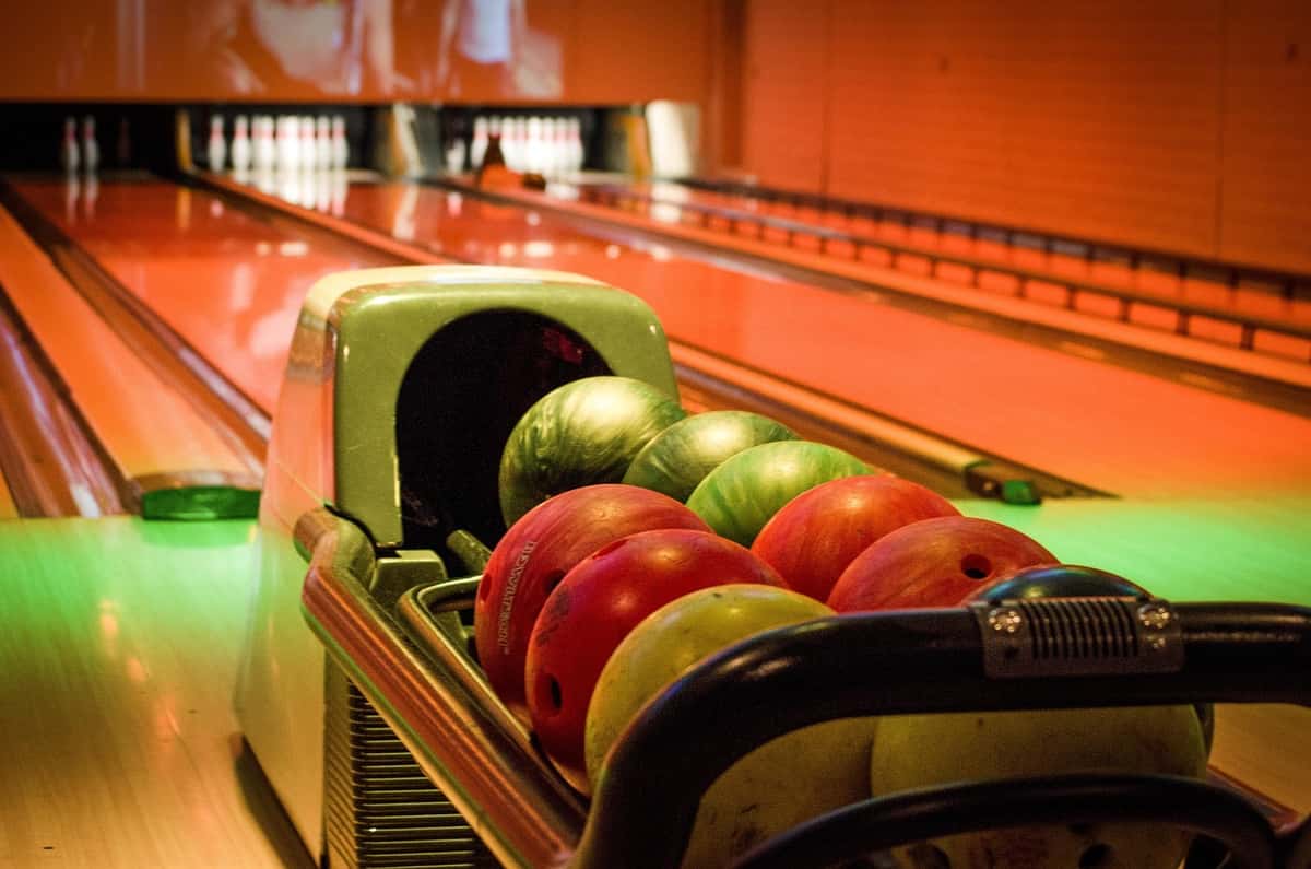 Rack of colorful bowling balls with glowing lanes in the background, leading to pins at the end of the alley.