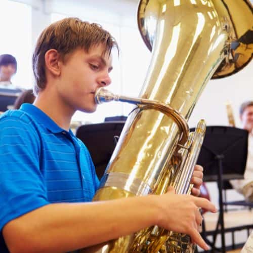 Teenage boy in a blue polo shirt plays a large brass tuba in a bright music classroom with other musicians in the background.