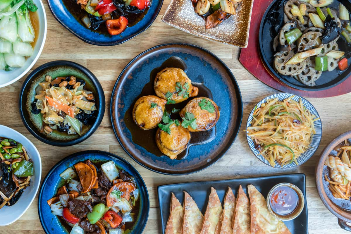 Assorted Thai dishes served on a wooden table including fried dumplings, stir-fried vegetables, noodles, and crispy wontons with sauce.