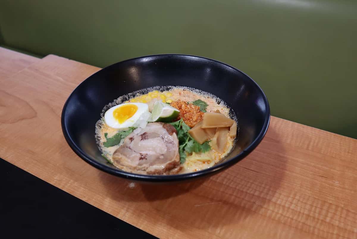 Bowl of ramen with soft-boiled egg, pork, bamboo shoots, corn, lime, and chili paste in a creamy broth