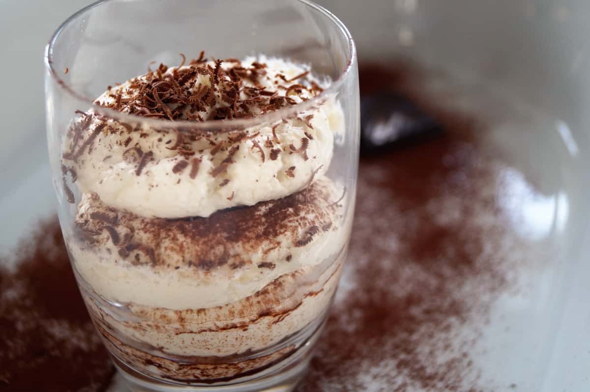 Glass cup filled with layered tiramisu, topped with whipped cream and chocolate shavings, surrounded by cocoa powder dusting on the surface.