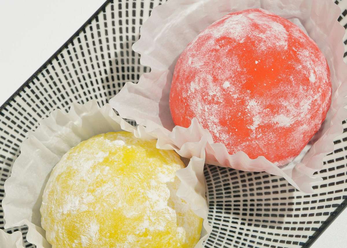 Colorful mochi ice cream balls in paper cups on a patterned tray, one red and one yellow, dusted with powdered sugar.