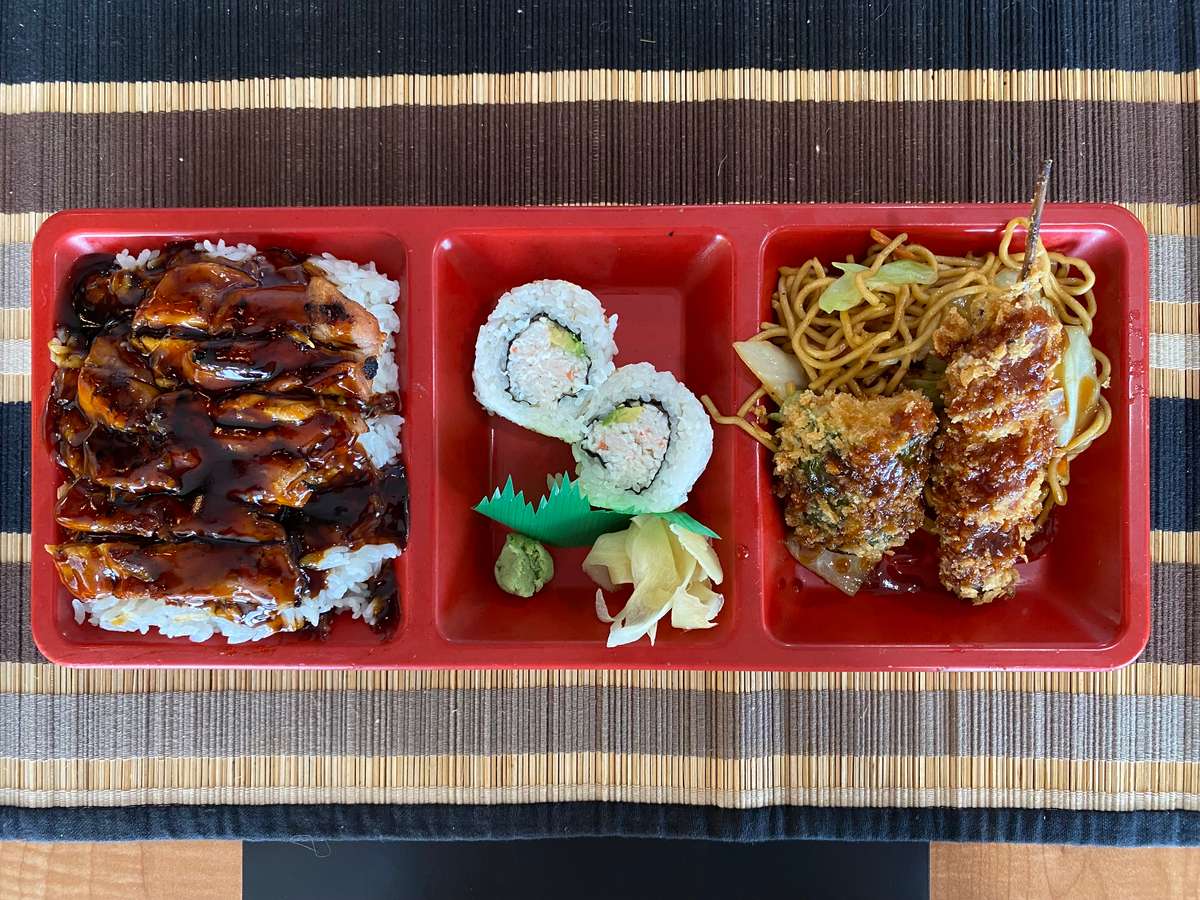 Japanese bento box with teriyaki chicken over rice, sushi rolls with wasabi and ginger, and fried skewers with noodles.